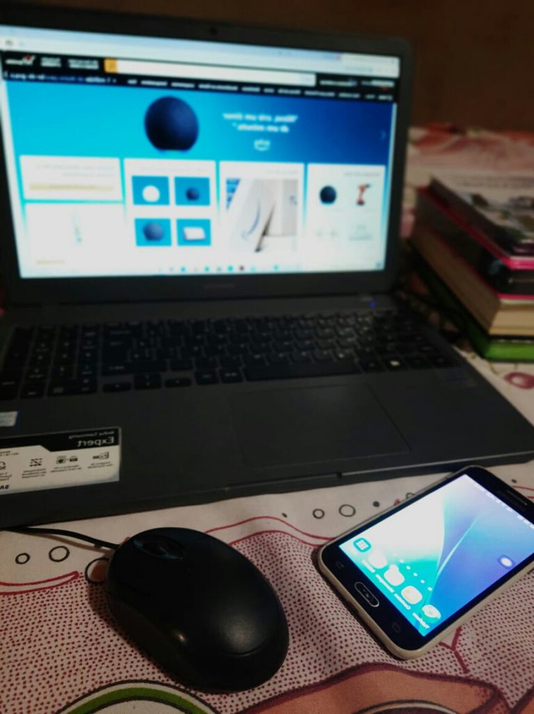 Tela de computador, mouse e celular estão em cima de uma mesa. Ao fundo livros e material e papelaria.