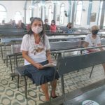 senhora sentada em banco de madeira da igreja, usa máscara, camiseta e saia. Ao fundo há mais alguns fiéis.