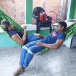 Casal deitado na rede de frente um para o outro usam o celular.