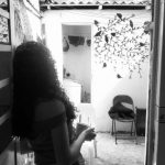 Foto em preto e branco mostra silhueta de jovem de cabelos compridos de costas com um copo na mão em frente há uma porta. A imagem tem um ar melancólico.
