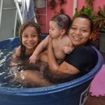 Duas crianças estão com a mãe em uma bacia improvisada como piscina. Eles estão sorrindo muito felizes.