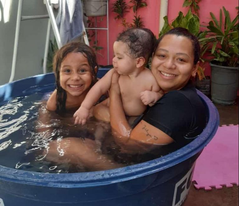 Duas crianças estão com a mãe em uma bacia improvisada como piscina. Eles estão sorrindo muito felizes.