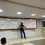 Professor em pé em frente ao quadro branco dando aula.