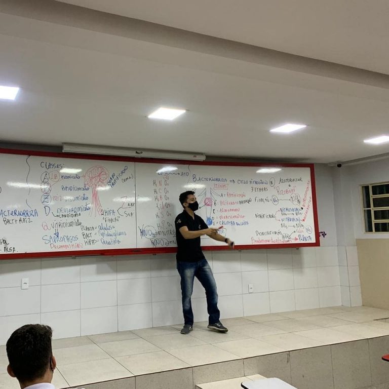 Professor em pé em frente ao quadro branco dando aula.