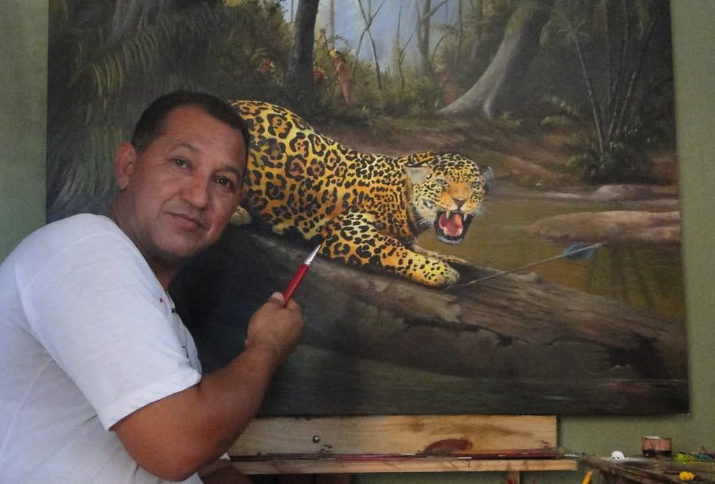 Ton Neves posa ao lado de uma pintura de uma onça-pintada em meio à floresta, segurando um pincel diante da obra em seu ateliê.
