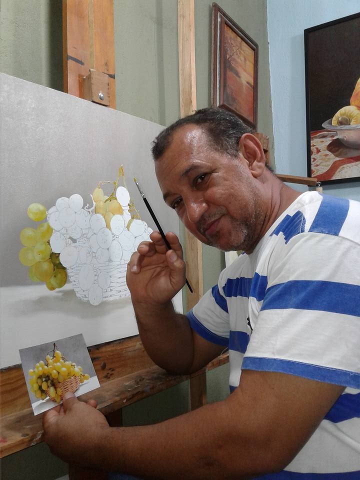 Ton Neves segura um pincel e uma fotografia de referência enquanto trabalha na pintura de um cacho de uvas em seu ateliê