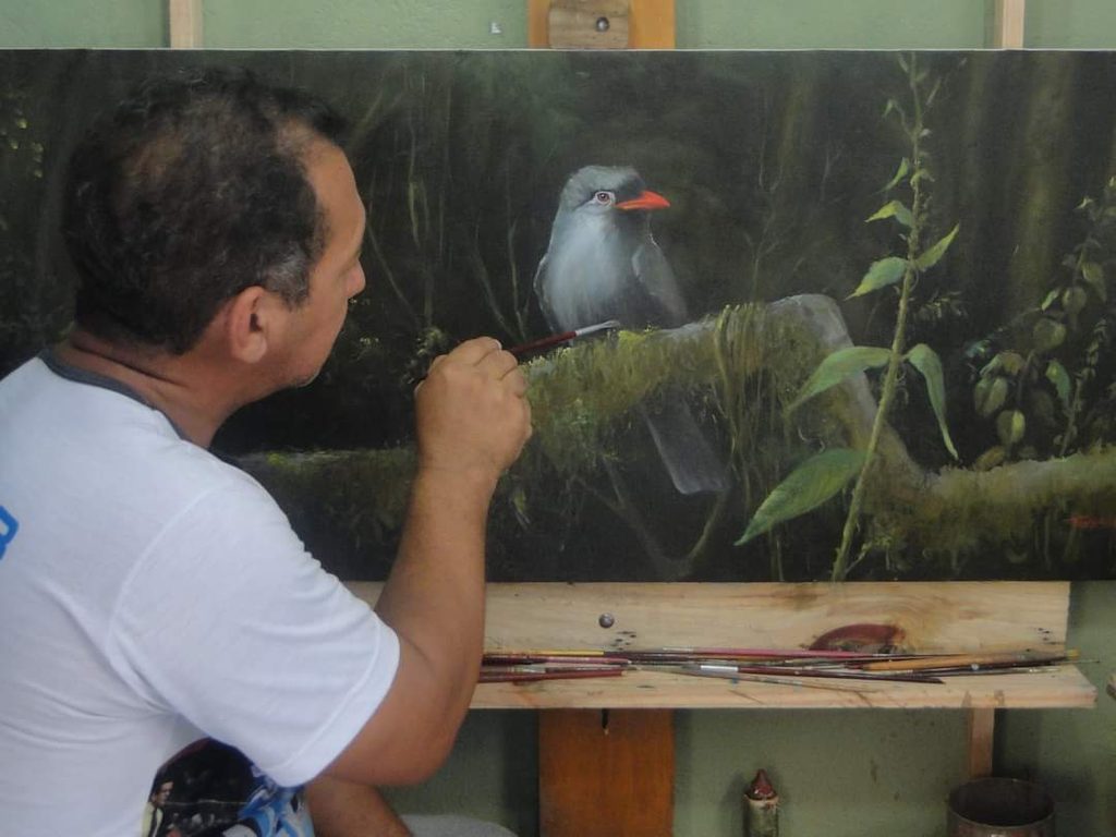 Ton Neves trabalha na pintura de um pássaro conhecido como “Bico de Brasa”, retratado em meio à vegetação, com detalhes que representam a fauna e a natureza maranhense.