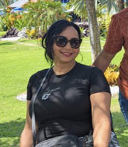 Uma mulher posa sentada em uma área verde, usando óculos escuros, camiseta preta e calça camuflada. Ela sorri para a foto, com uma bolsa preta atravessada no corpo. Ao fundo, há palmeiras, jardins e uma área de lazer iluminada pelo sol.